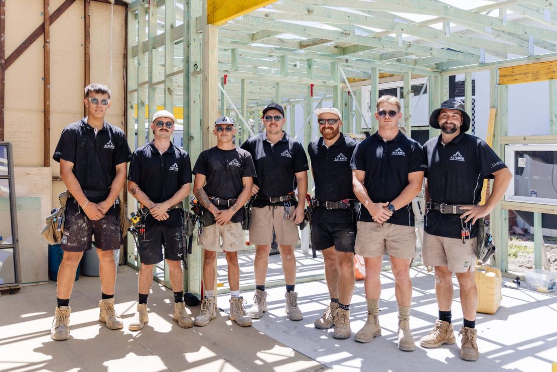 A group of seven people in dark shirts and tan shorts stand in a line on a wooden construction site.