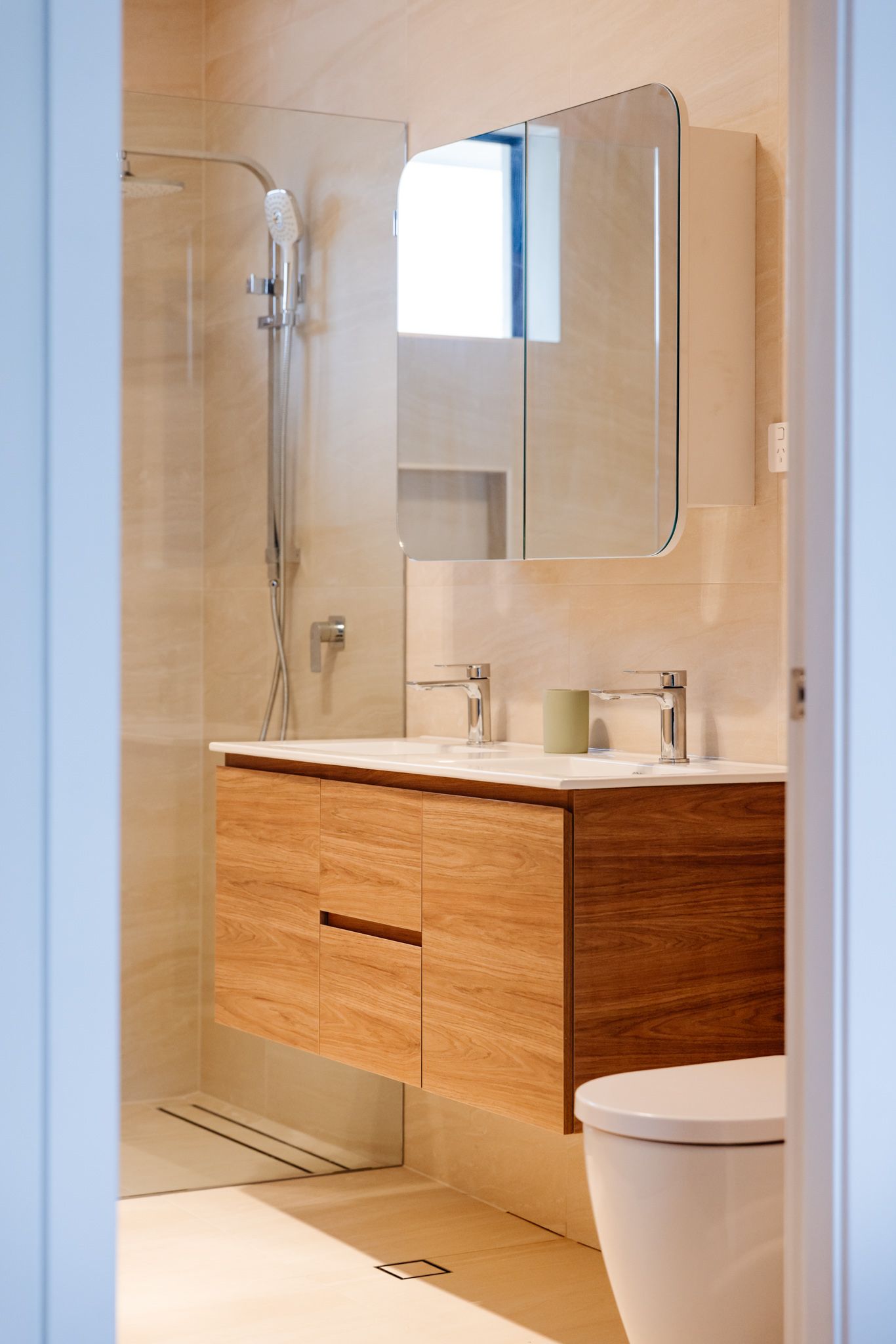 A modern bathroom with a light wood floating vanity, double sinks, a mirrored medicine cabinet, and a glass-enclosed shower.