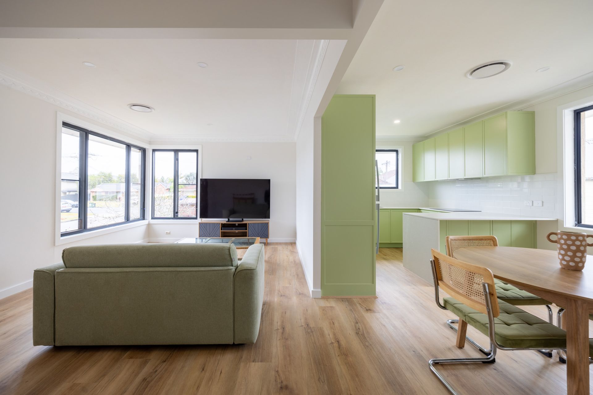 A modern open-plan home featuring a sage green sofa, a television area, and a kitchen with light green cabinets.
