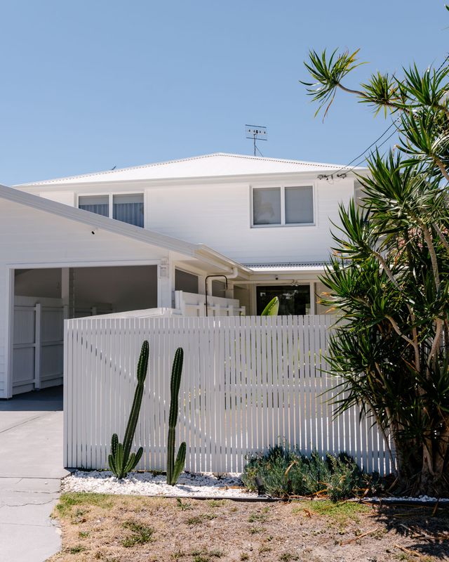 A two-story white house with a matching picket fence, a carport, and green plants in the front yard under a blue sky.