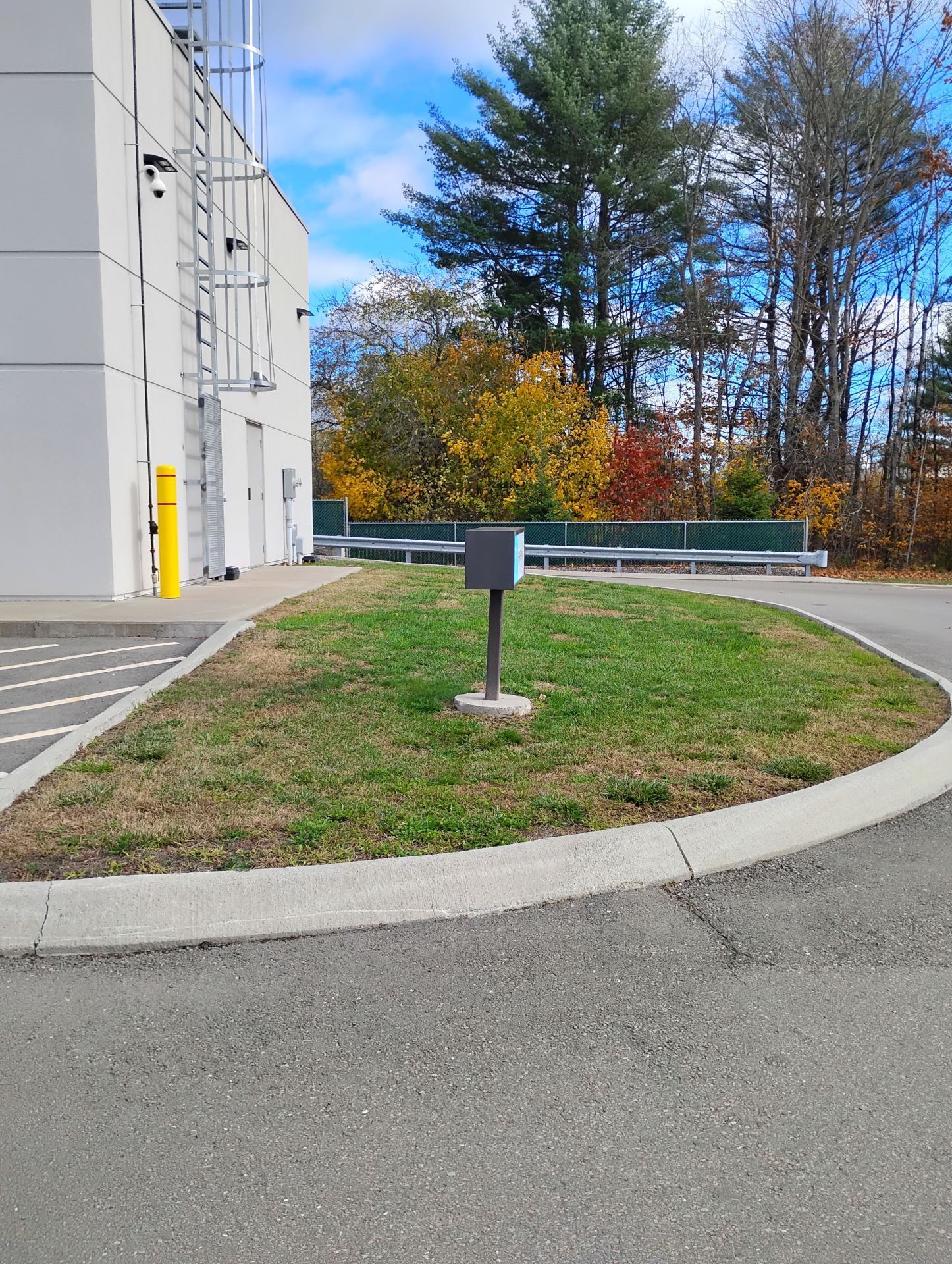Grassy median with a metal box and curb next to a building and paved area. Fall foliage in the background.