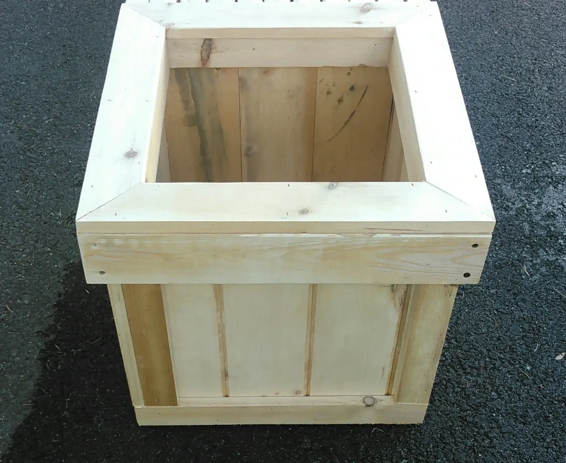 Wooden square planter box, unpainted with a framed top, set outside on dark pavement.