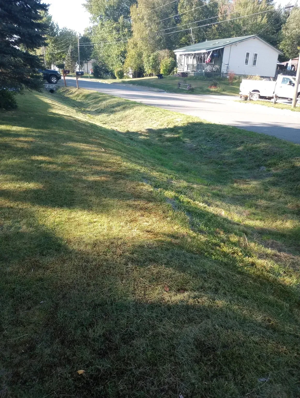 Grassy area slopes down to a residential street with a white house and trees in the background.