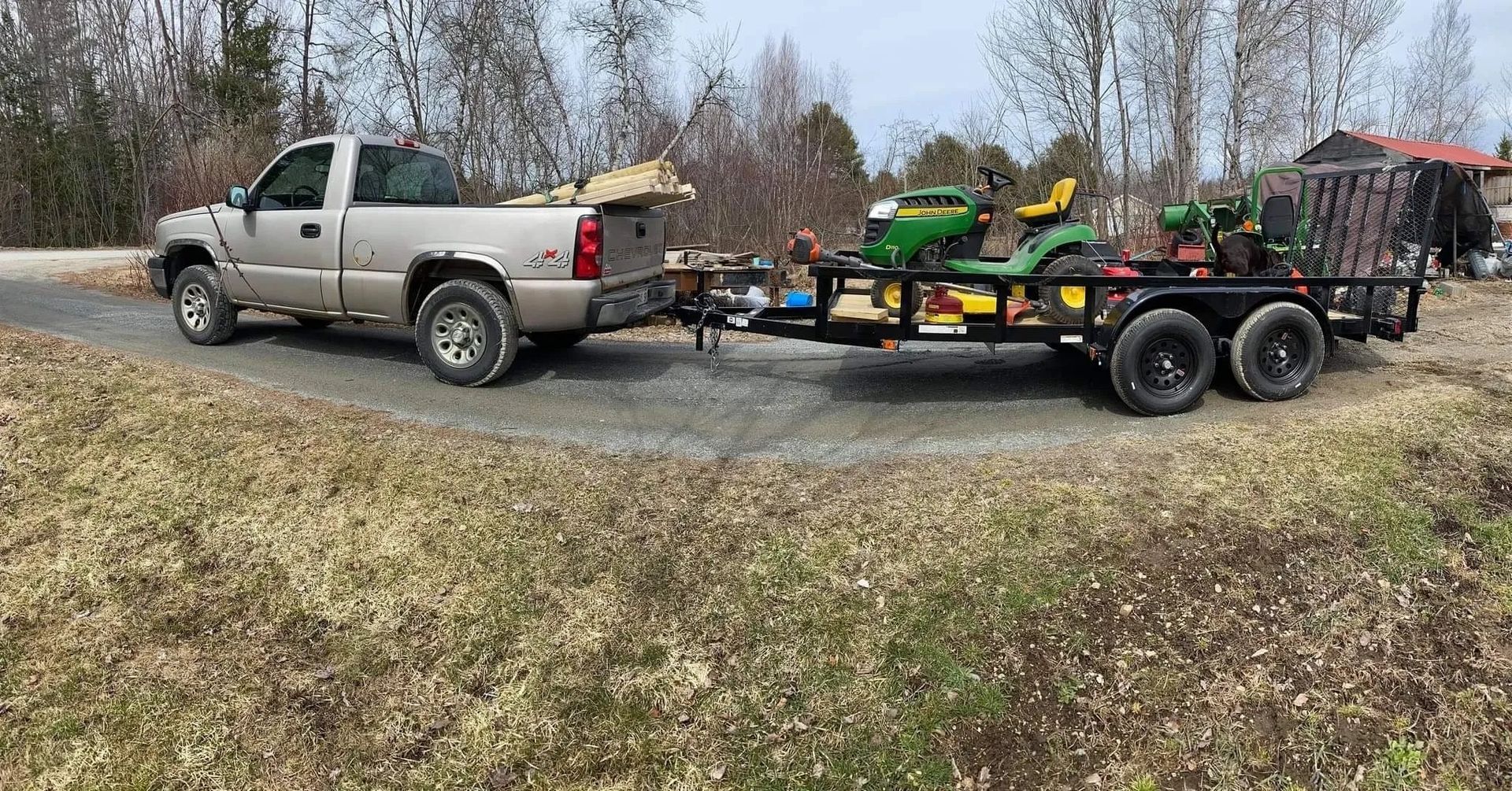 Tan pickup truck towing a trailer with a green riding lawnmower and a snow blower on a dirt road.