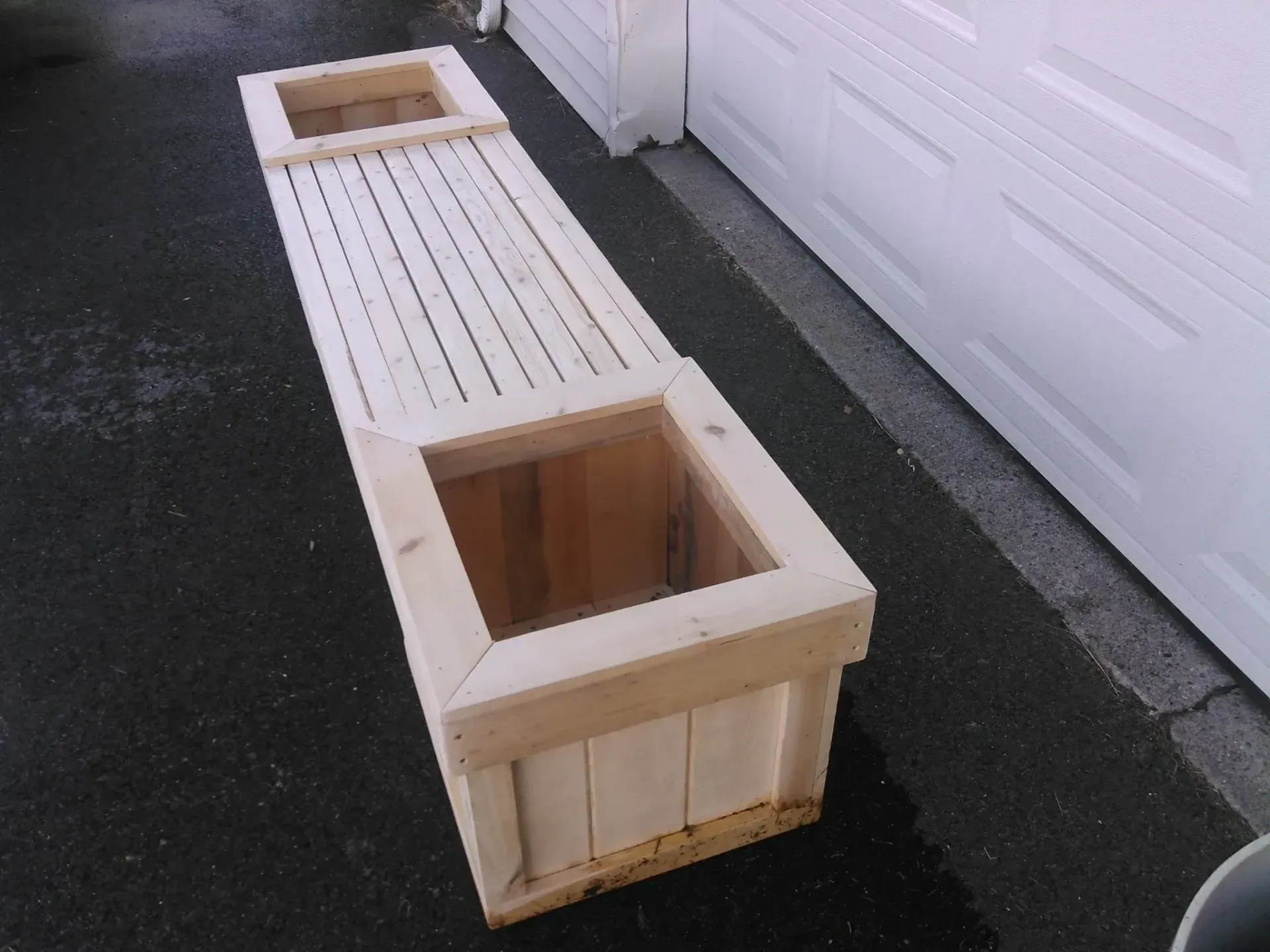 Wooden planter bench with two square planters, slatted seat, on asphalt next to a white garage door.