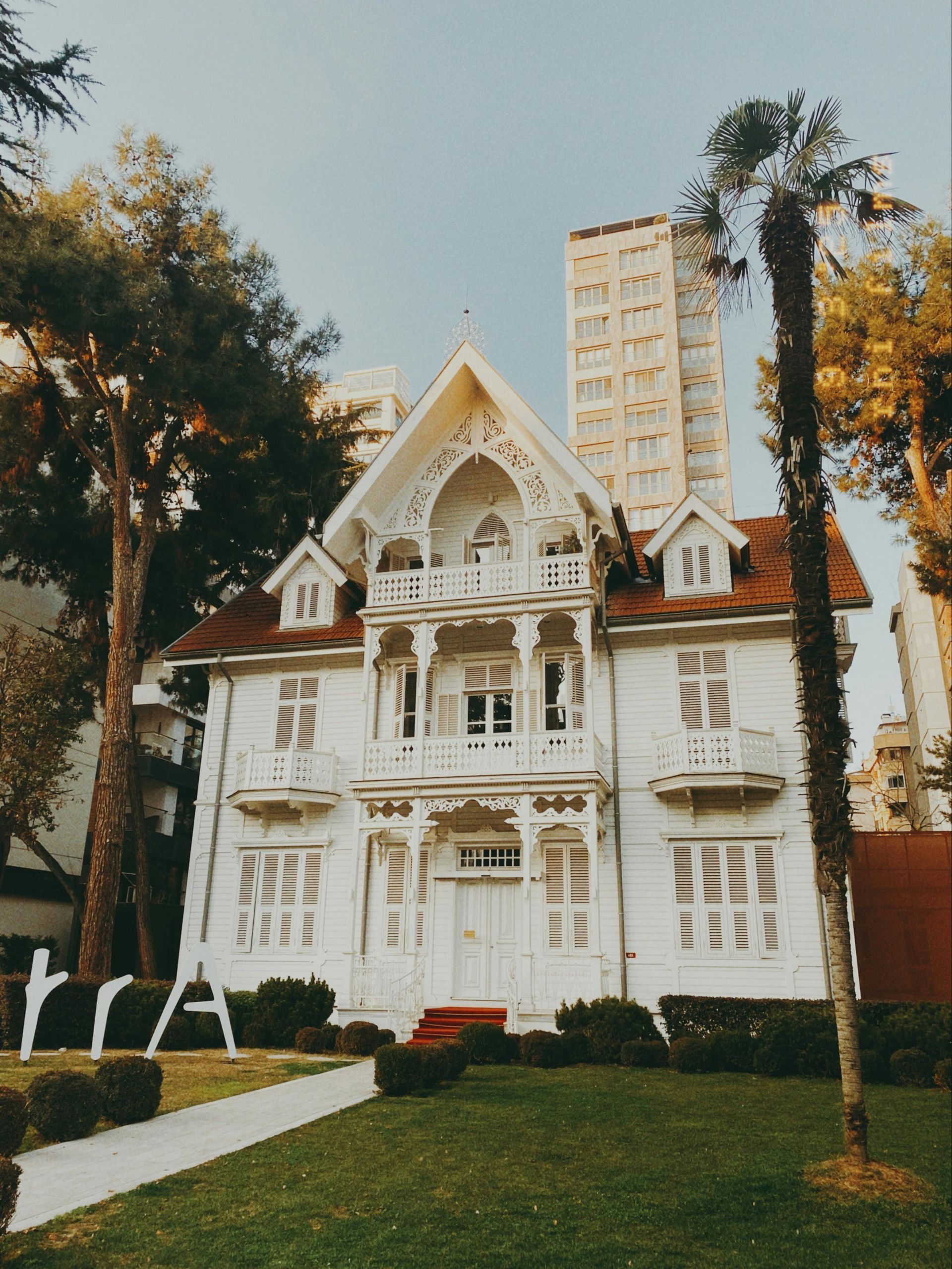 White Victorian house with ornate details and a red roof, trees, and buildings in the background.
