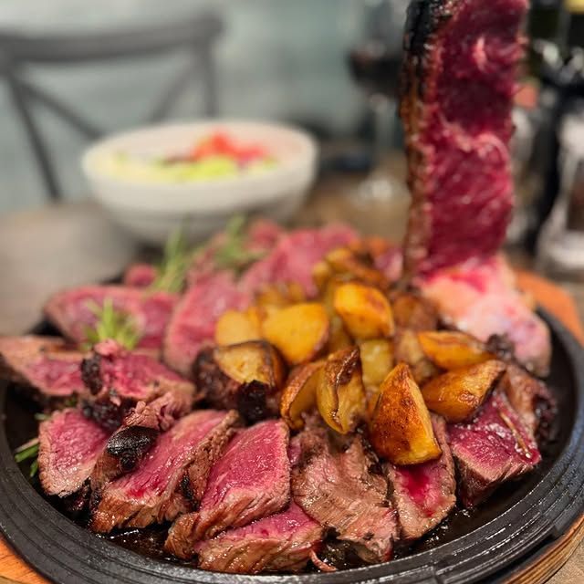 Piatto di bistecca a fette, cotta al sangue, con patate arrosto, servito su una piastra rovente, con una ciotola di contorno sullo sfondo.