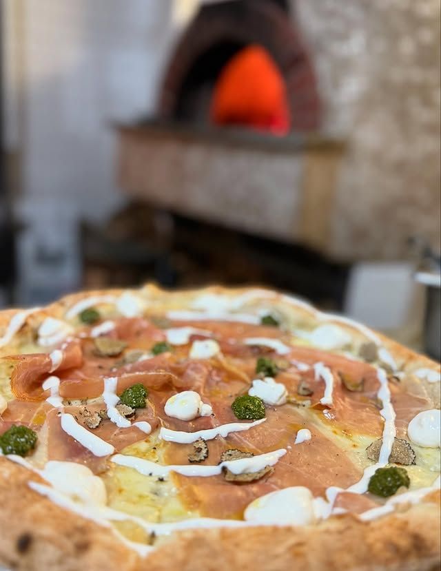 Primo piano di una pizza bianca con condimenti, posta davanti a un forno a legna sfocato.