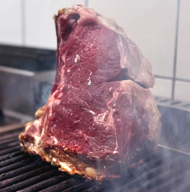 Una grossa bistecca cruda viene scottata sulla griglia, con il fumo che si alza in un ambiente di cottura all'aperto.