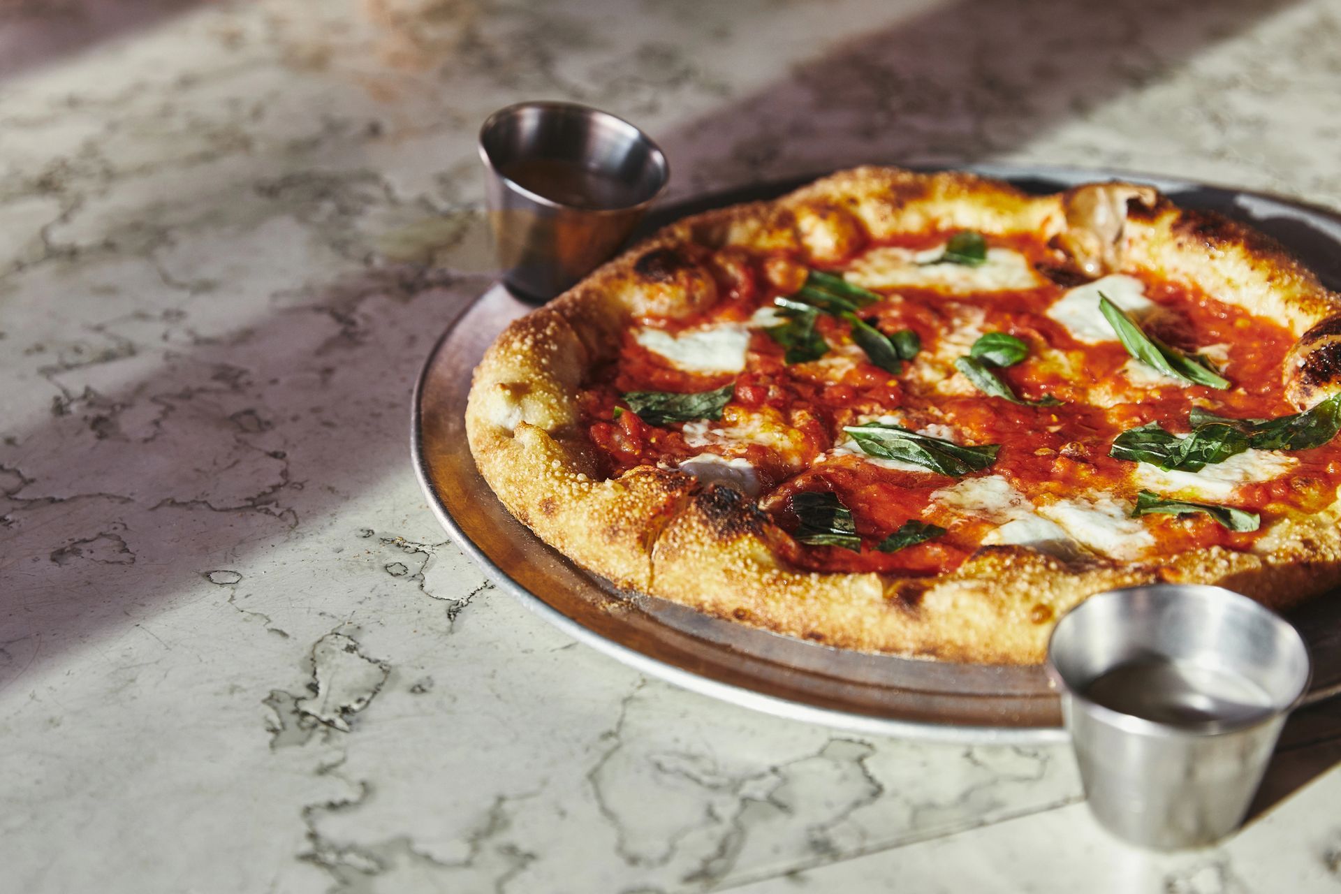 Pizza cotta nel forno a legna su un vassoio di metallo con due tazze d'acciaio su un tavolo di marmo.