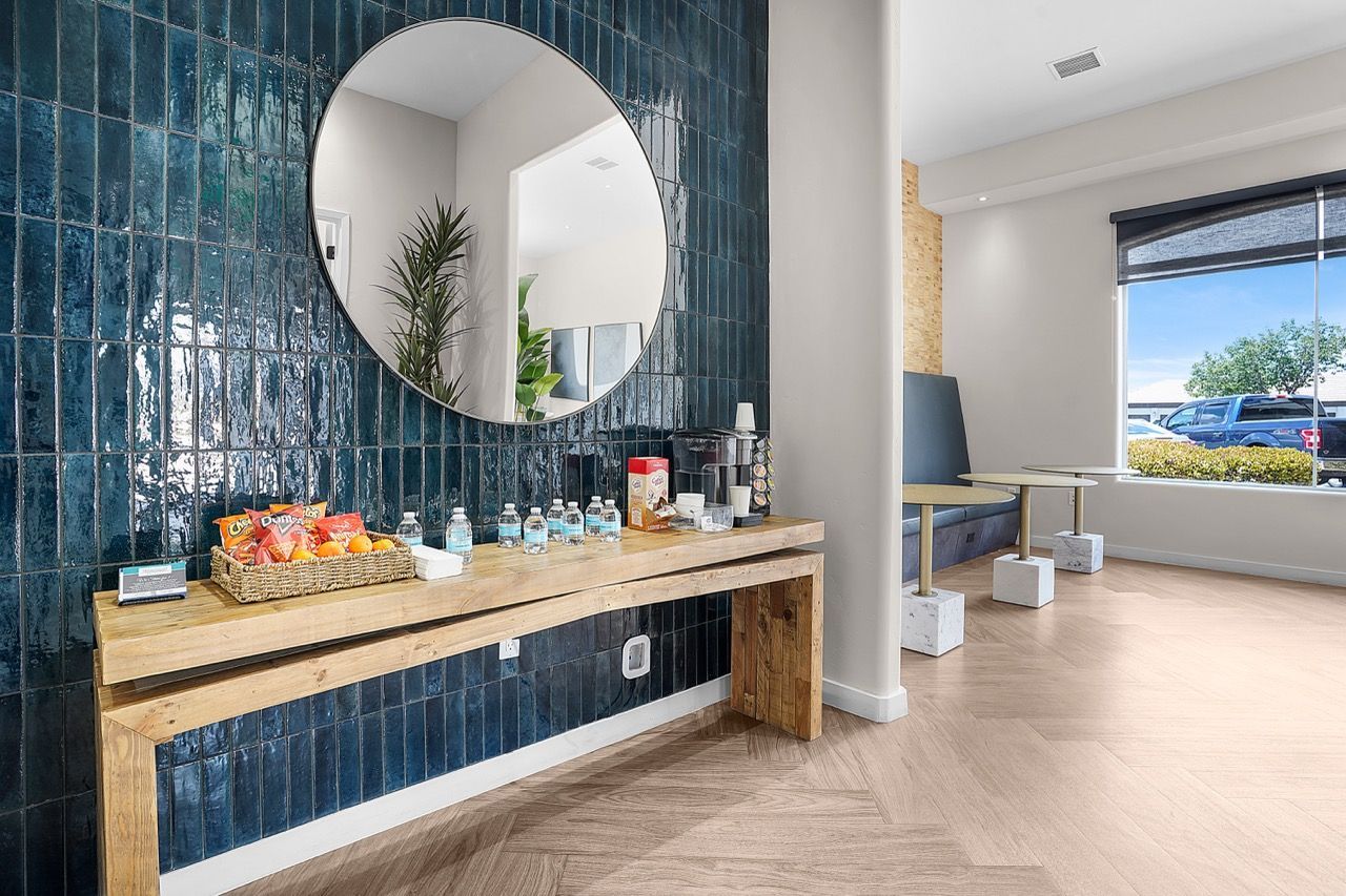 Lobby area with a wooden console, round mirror, water bottles, snacks, and seating by a window at Broadstone Heights in Albuquerque, NM.