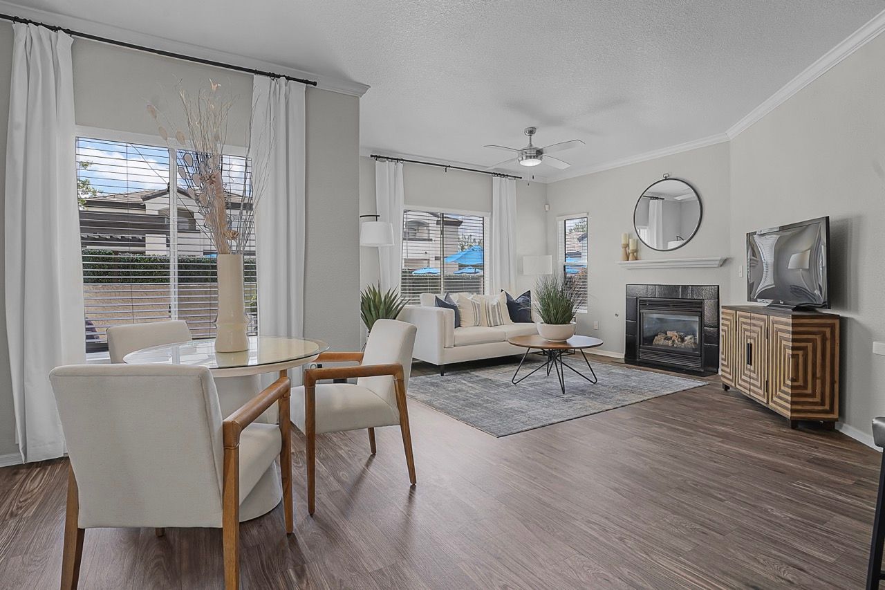 Open-concept living room with white sofa, round coffee table, dining set, fireplace, and large windows with white curtains at Broadstone Heights in Albuquerque, NM.