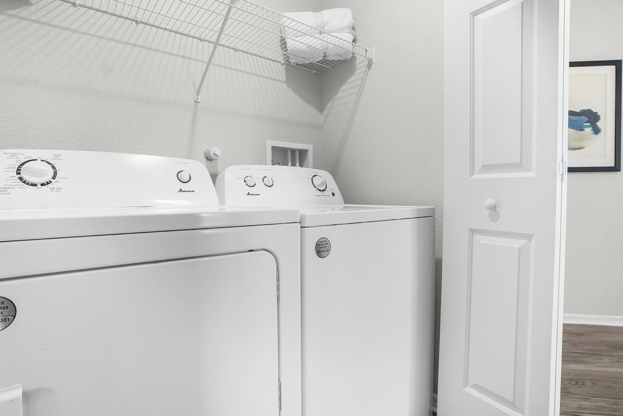 White washer and dryer in a small apartment laundry room with a wire shelf and an open door at Broadstone Heights in Albuquerque, NM.
