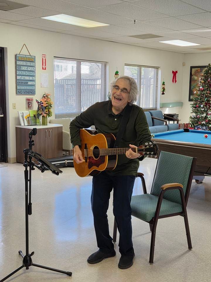 Primeplus volunteer playing guitar and singing indoors, by a pool table and Christmas tree. He's wearing glasses and smiling.