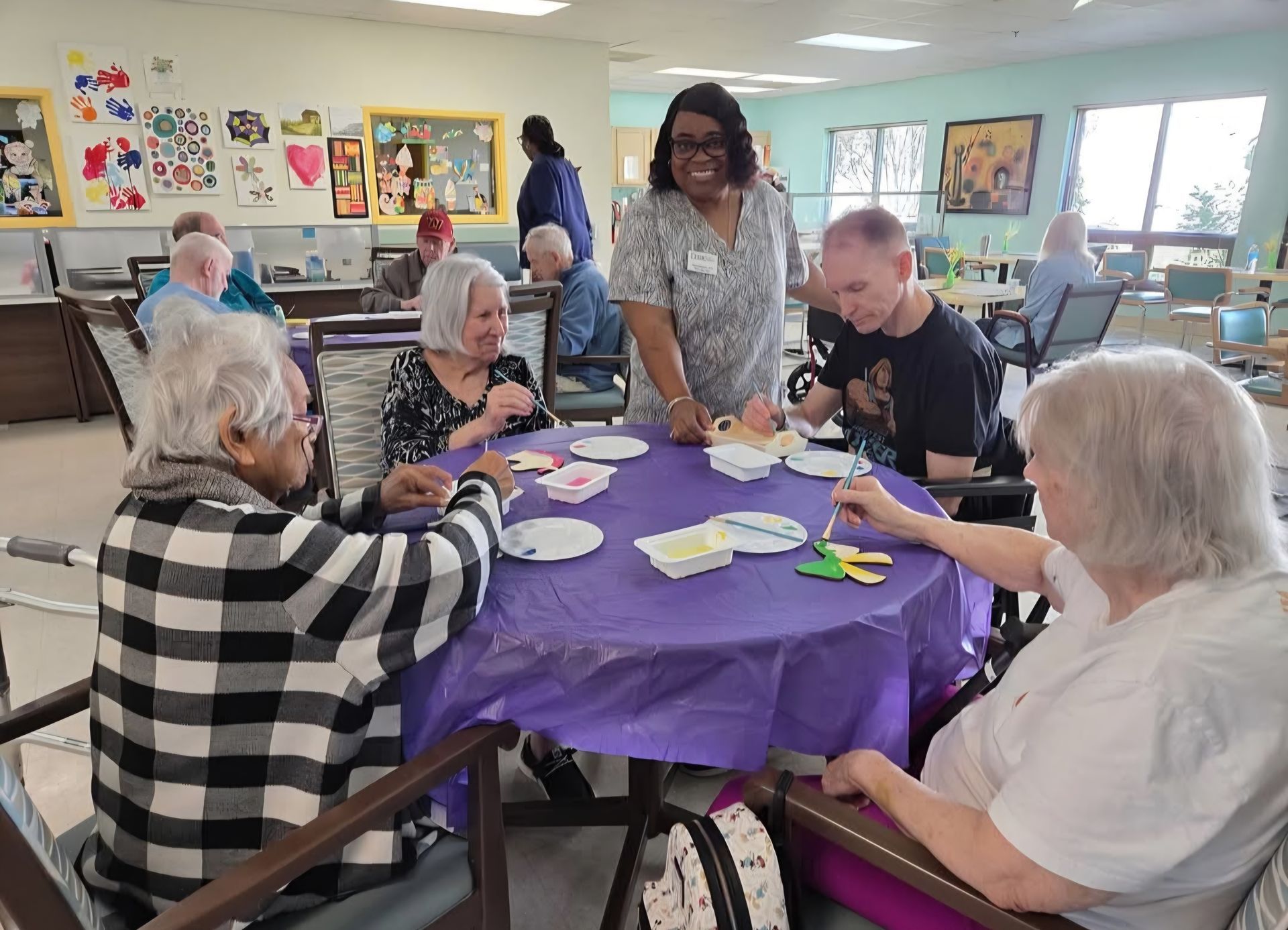 Primeplus Adult Day Care participants playing a game at a table in a brightly lit room, overseen by a staff member.
