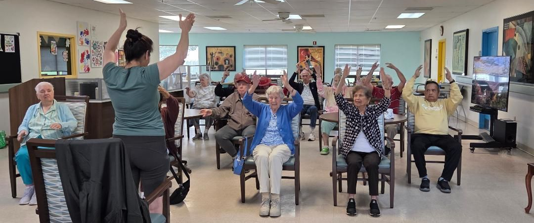 Primeplus Members exercise in a brightly lit room while sitting in chairs; a woman leads the activity.