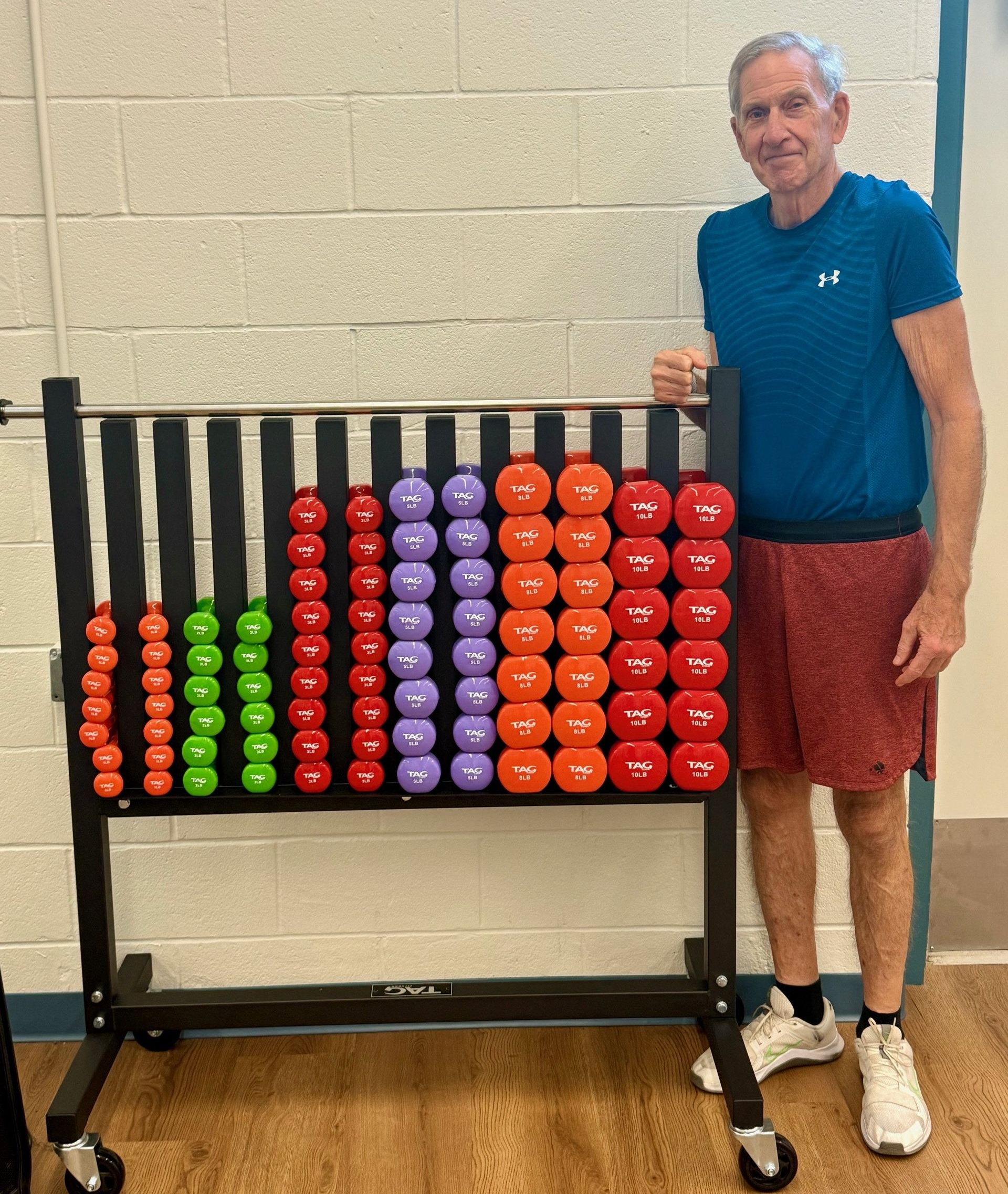 Primeplus Member stands beside a colorful exercise weight rack with wheels in a gym.