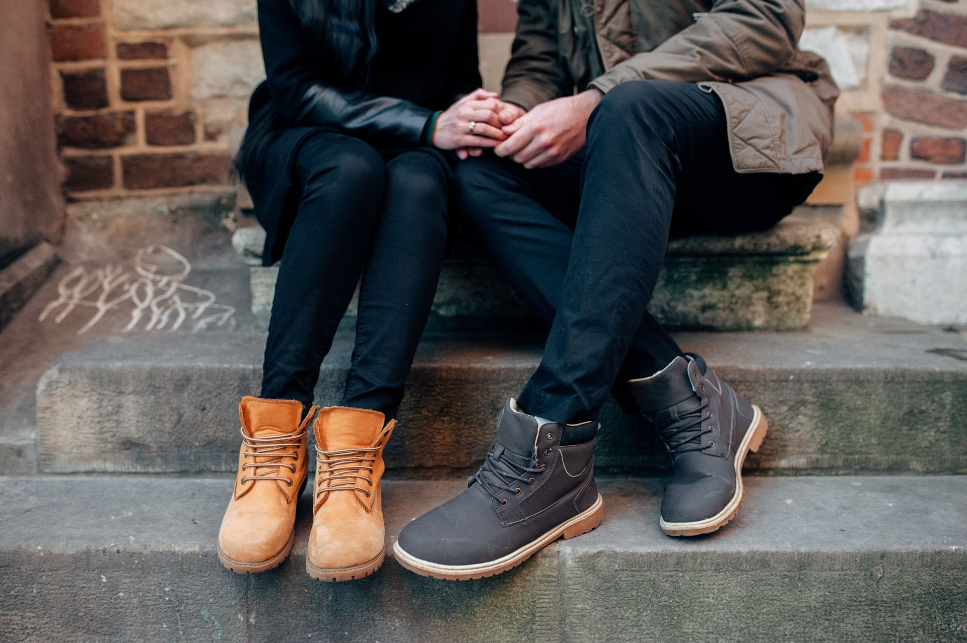 Two people holding hands, sitting on steps; one wears tan boots, the other gray.