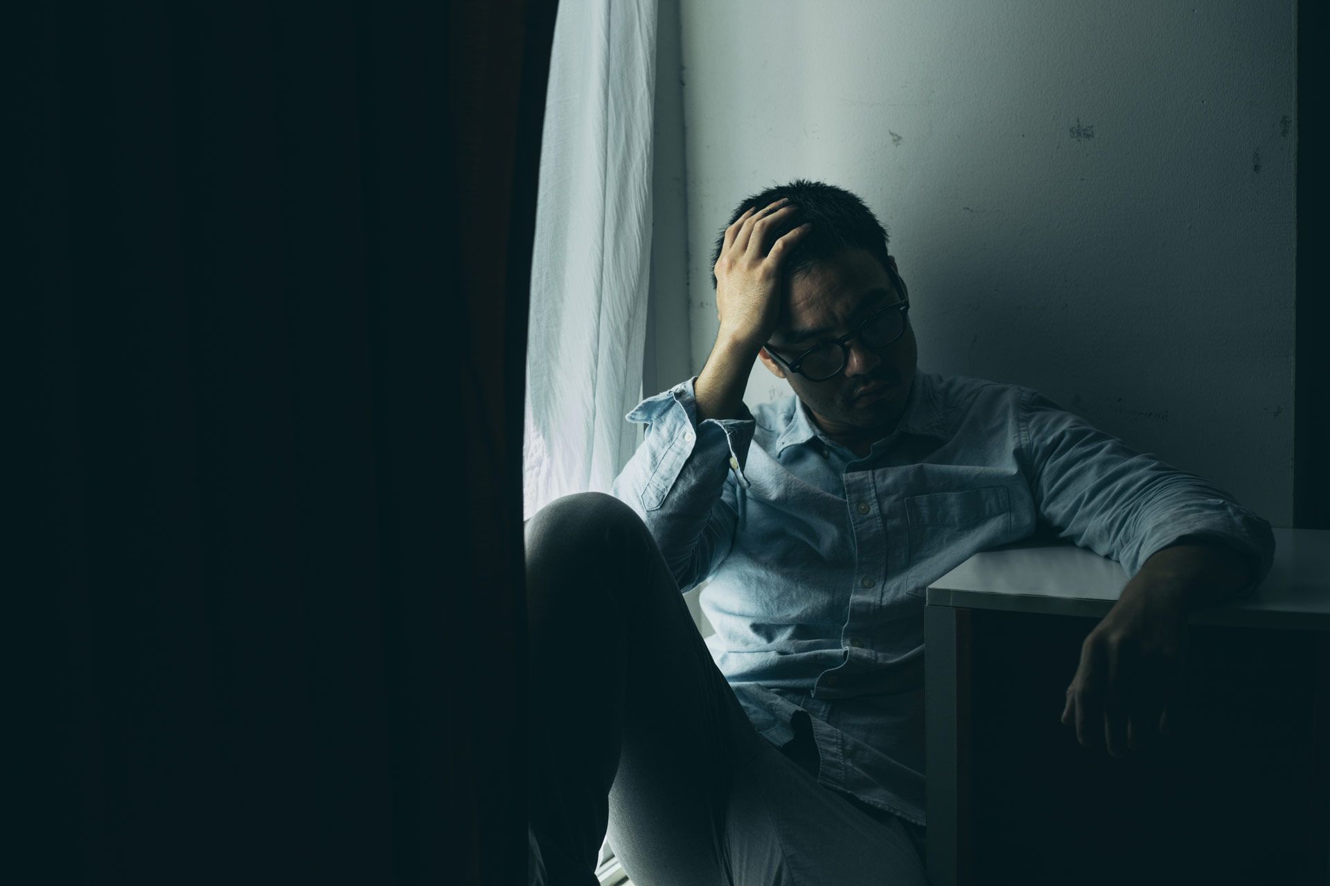 Man sits slumped against a wall by a window, hand on head, looking downcast.