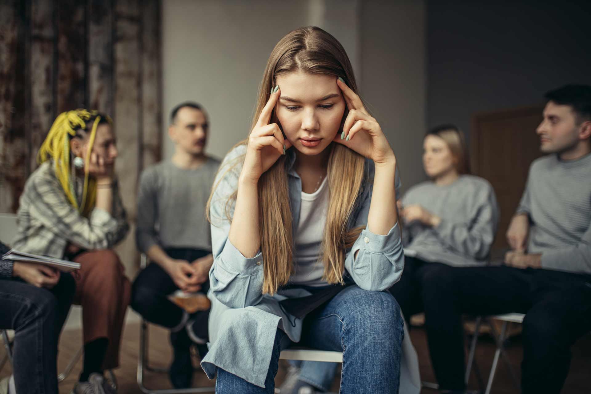 Woman with hands on head sits in group therapy session, looking distressed.