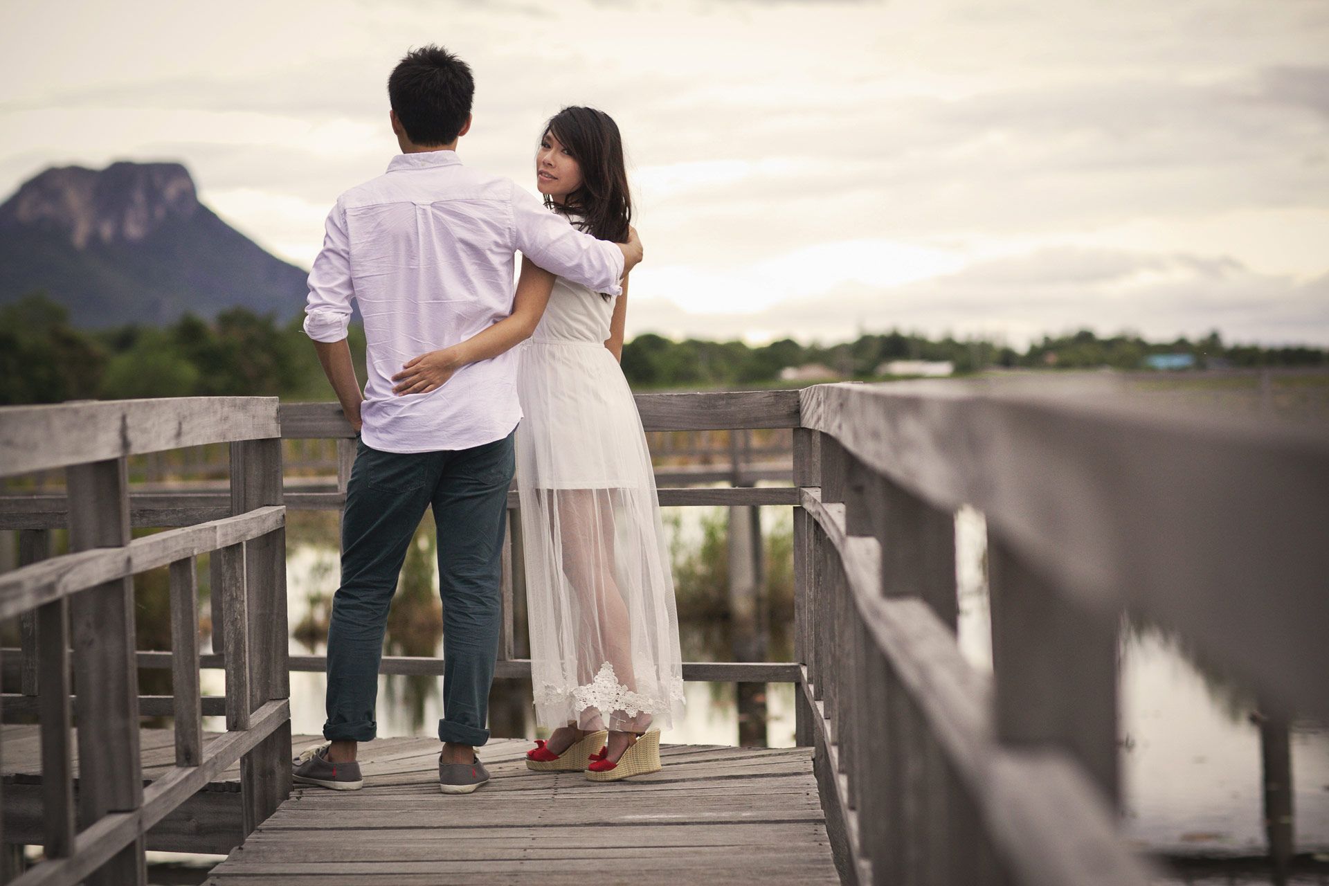 Couple embracing on a wooden bridge, looking at a mountain.