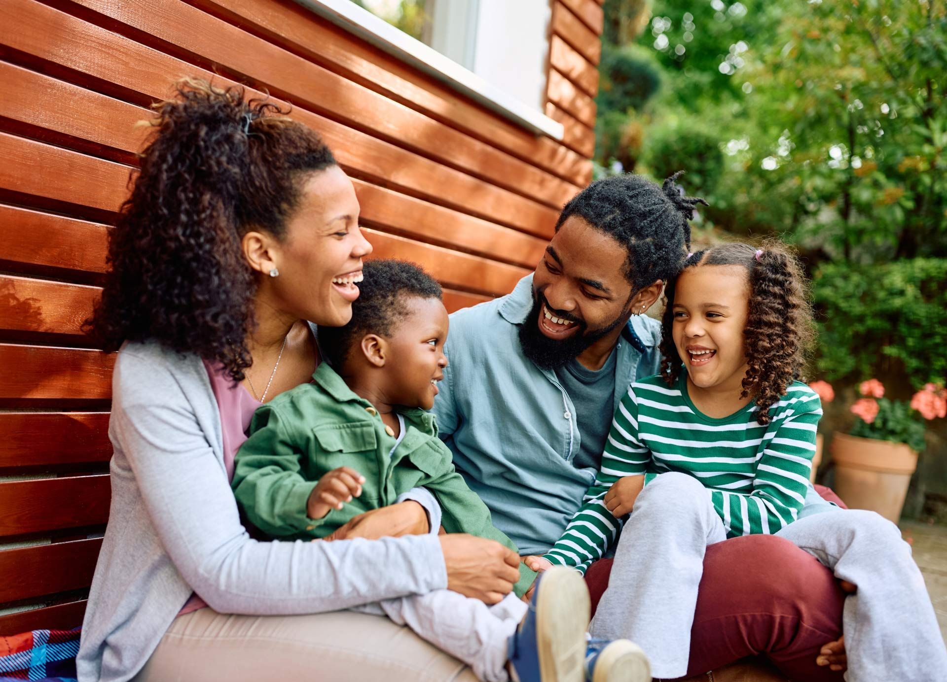 Family laughing together outdoors, sitting near a wooden wall, with green foliage in the background.