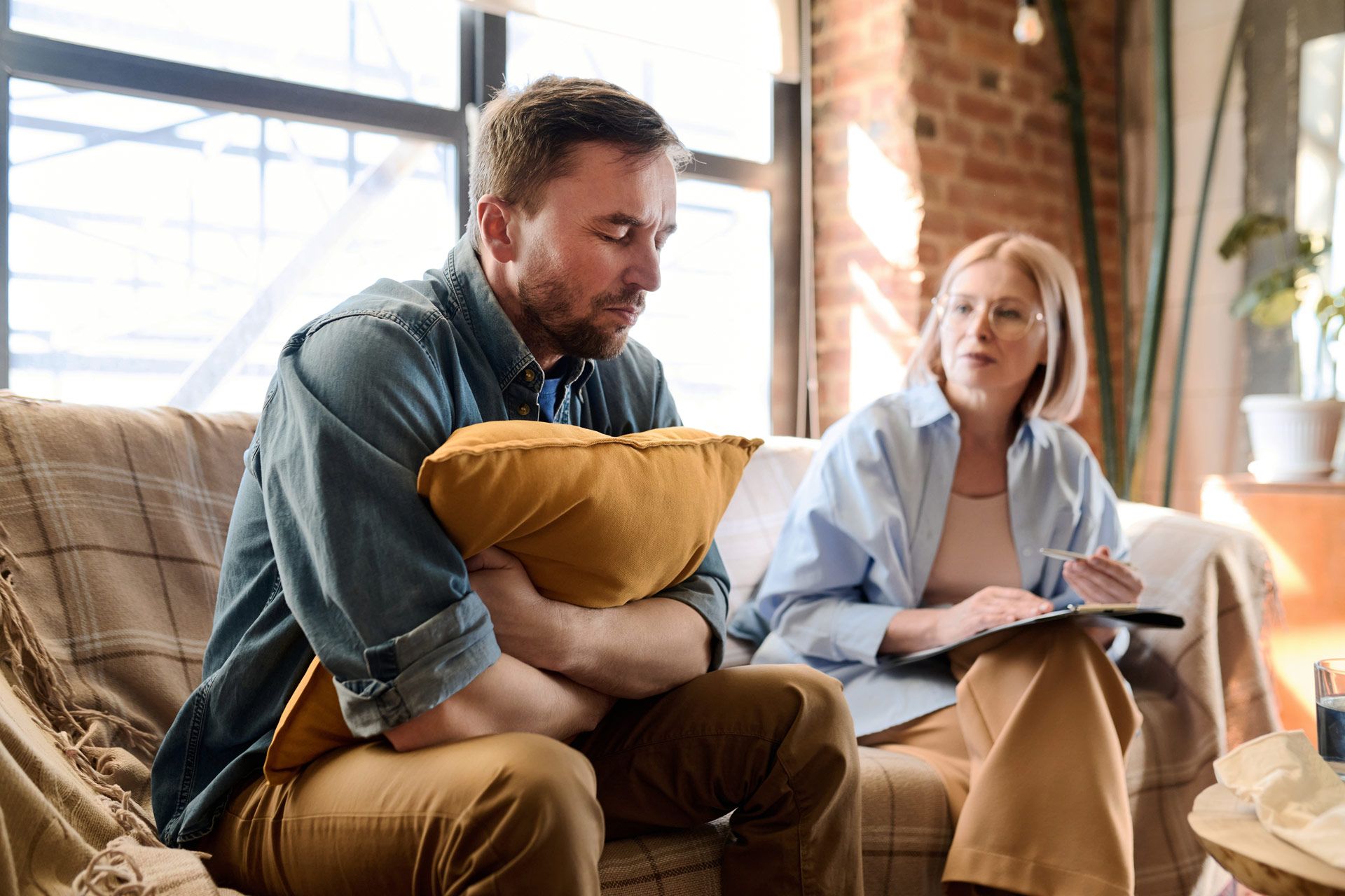 Man hugging pillow, talking with therapist on couch. Interior setting, brick wall, natural light.