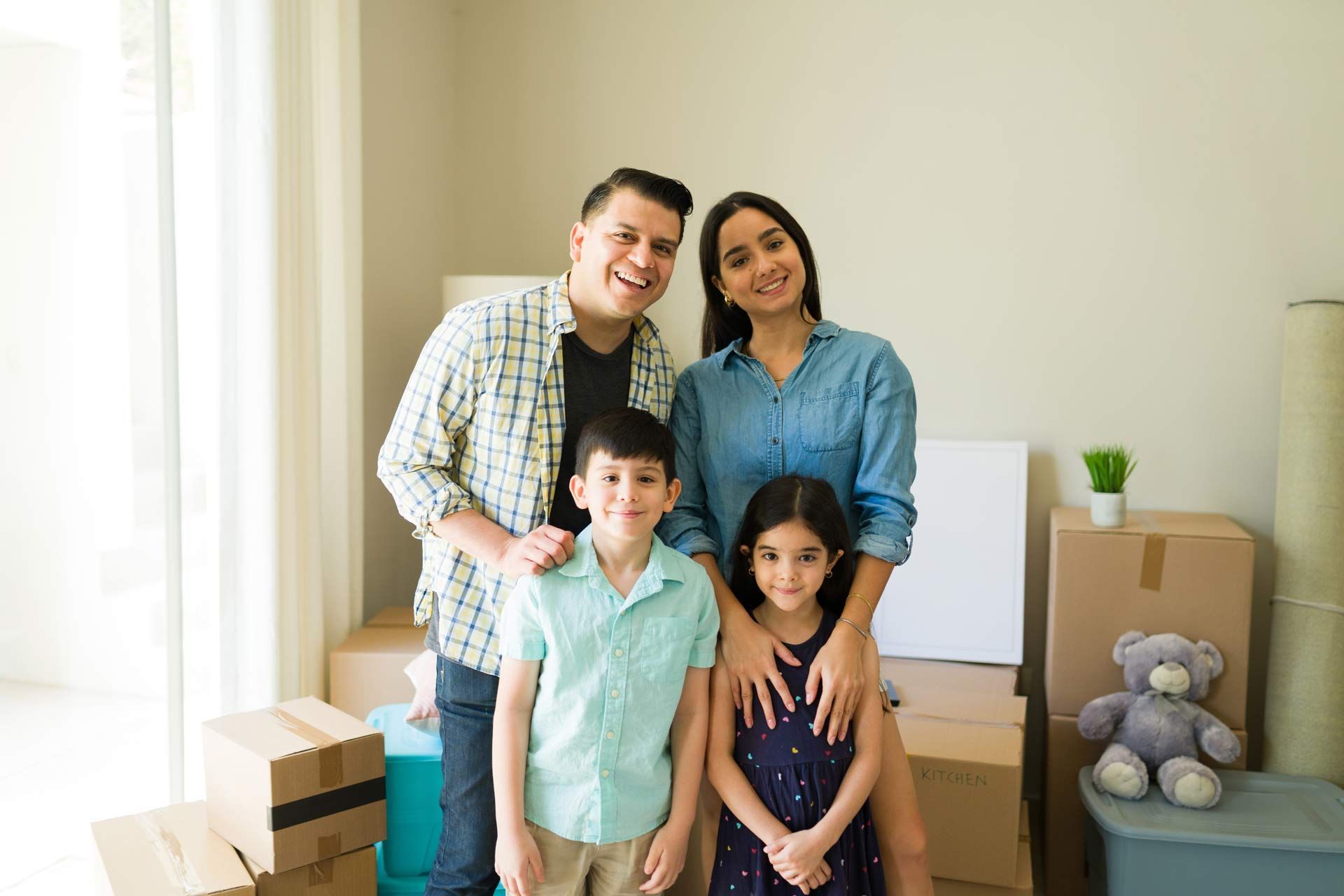 Family of four smiling in a new home, surrounded by moving boxes.