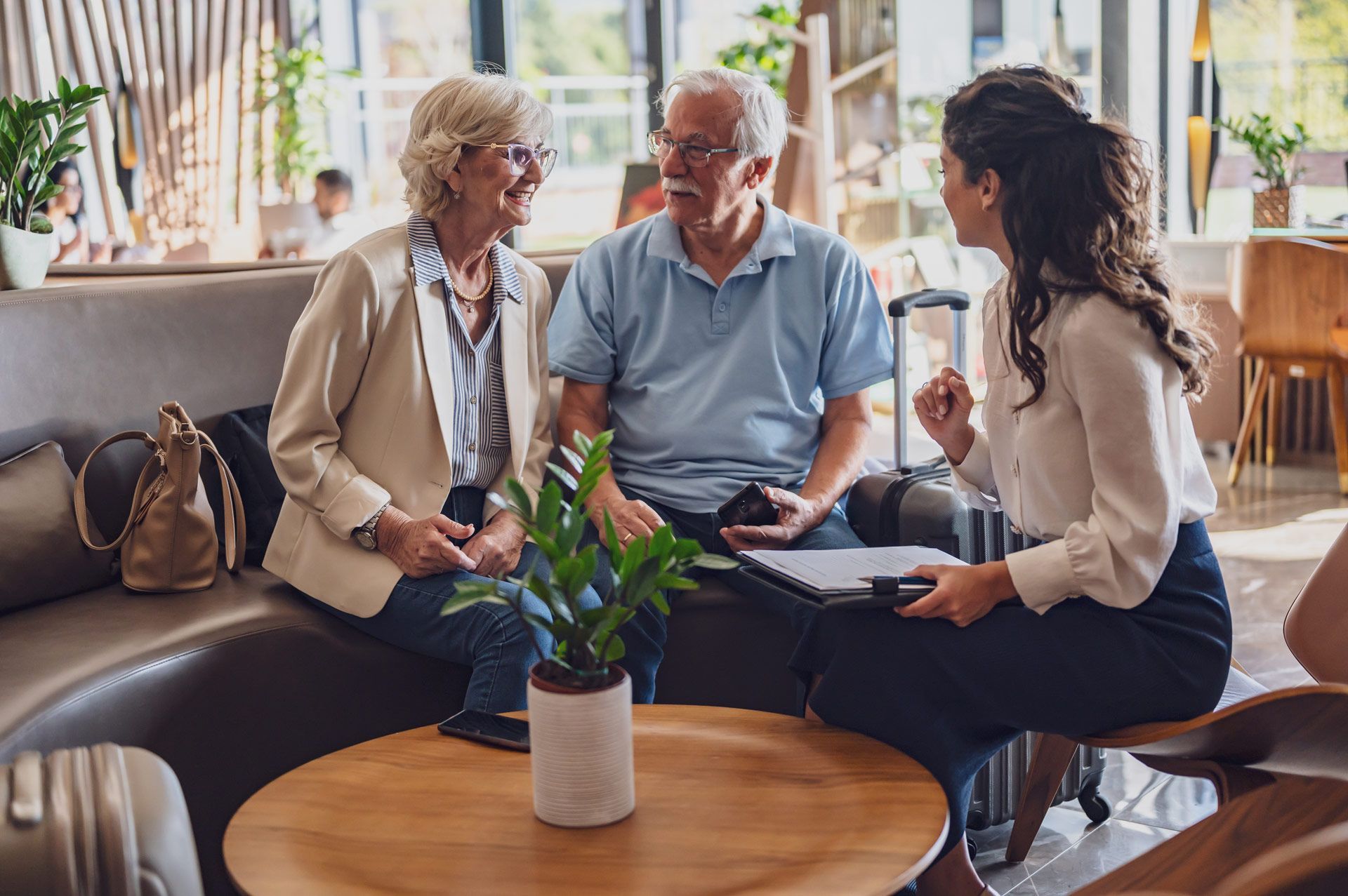 Elderly couple talking to a travel agent, sitting near a small plant and suitcases.