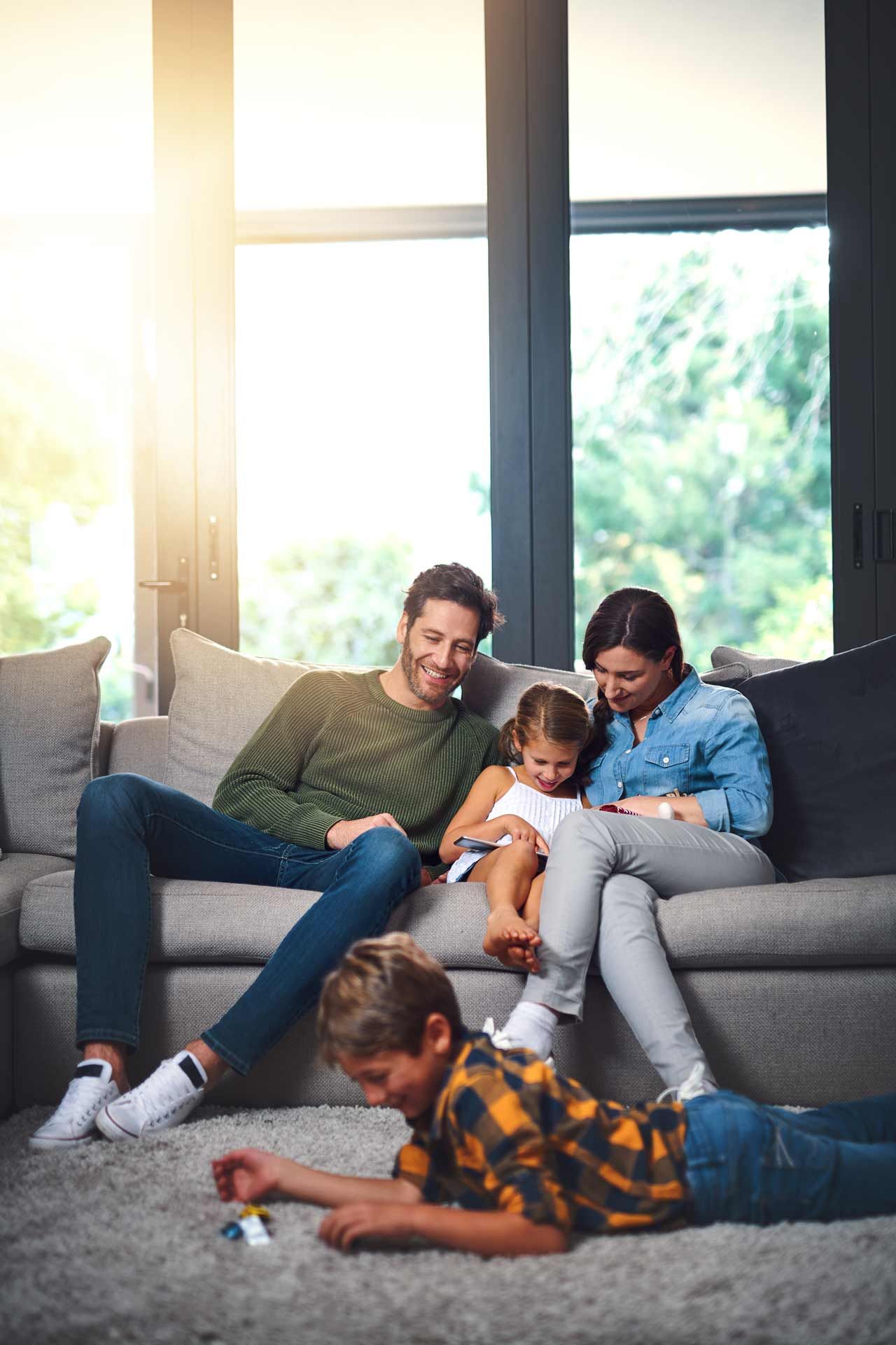 Family on a sofa, looking at a device, son on the floor with toys. Bright, modern living room.