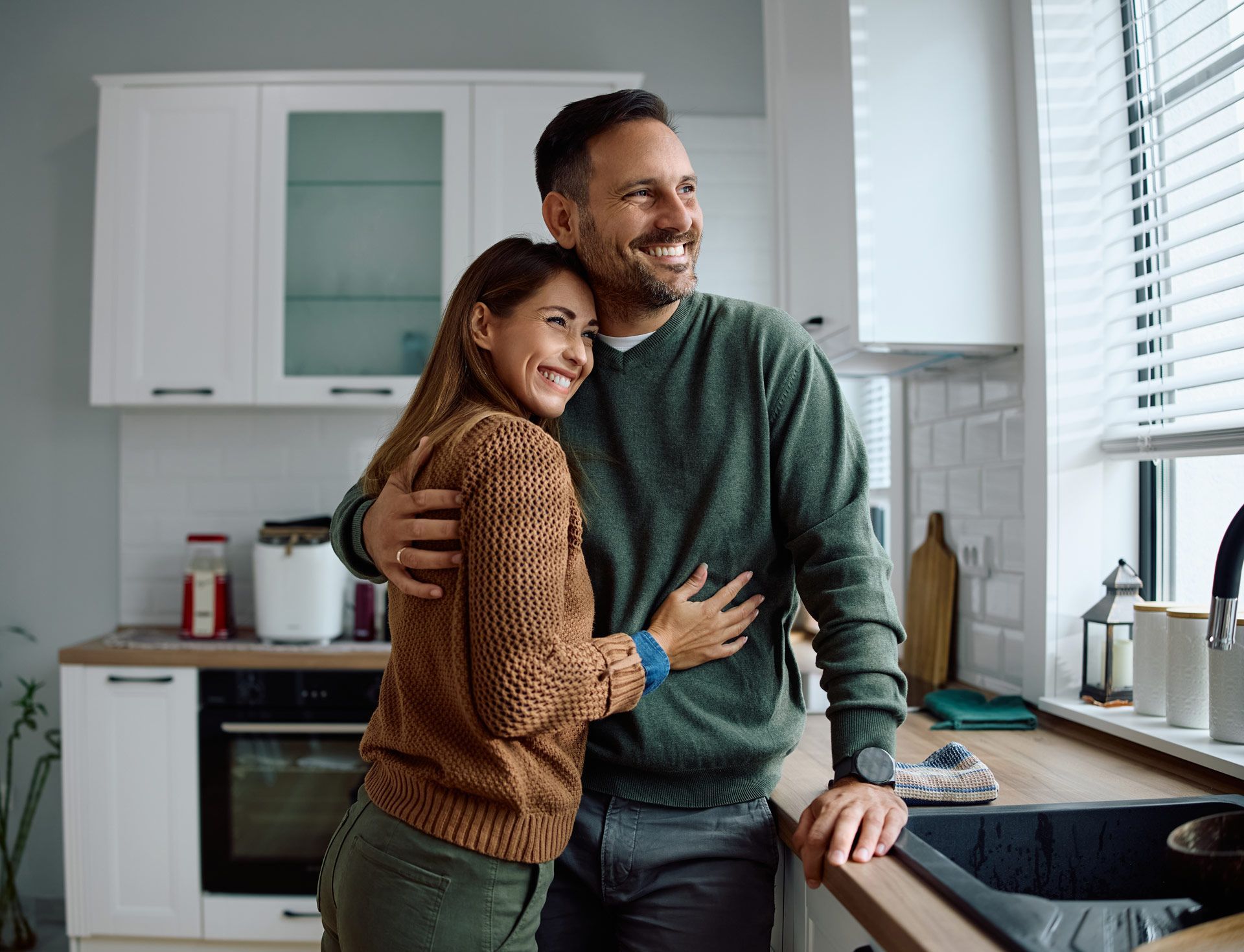 Couple embracing in a kitchen, smiling and looking out a window.