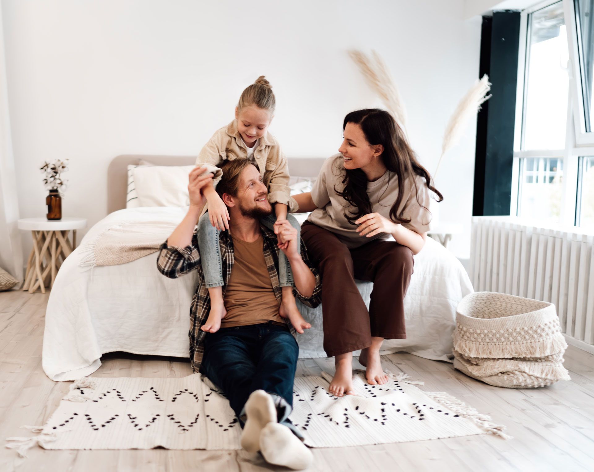 Family in bedroom; father with child on shoulders, mother sits nearby smiling.