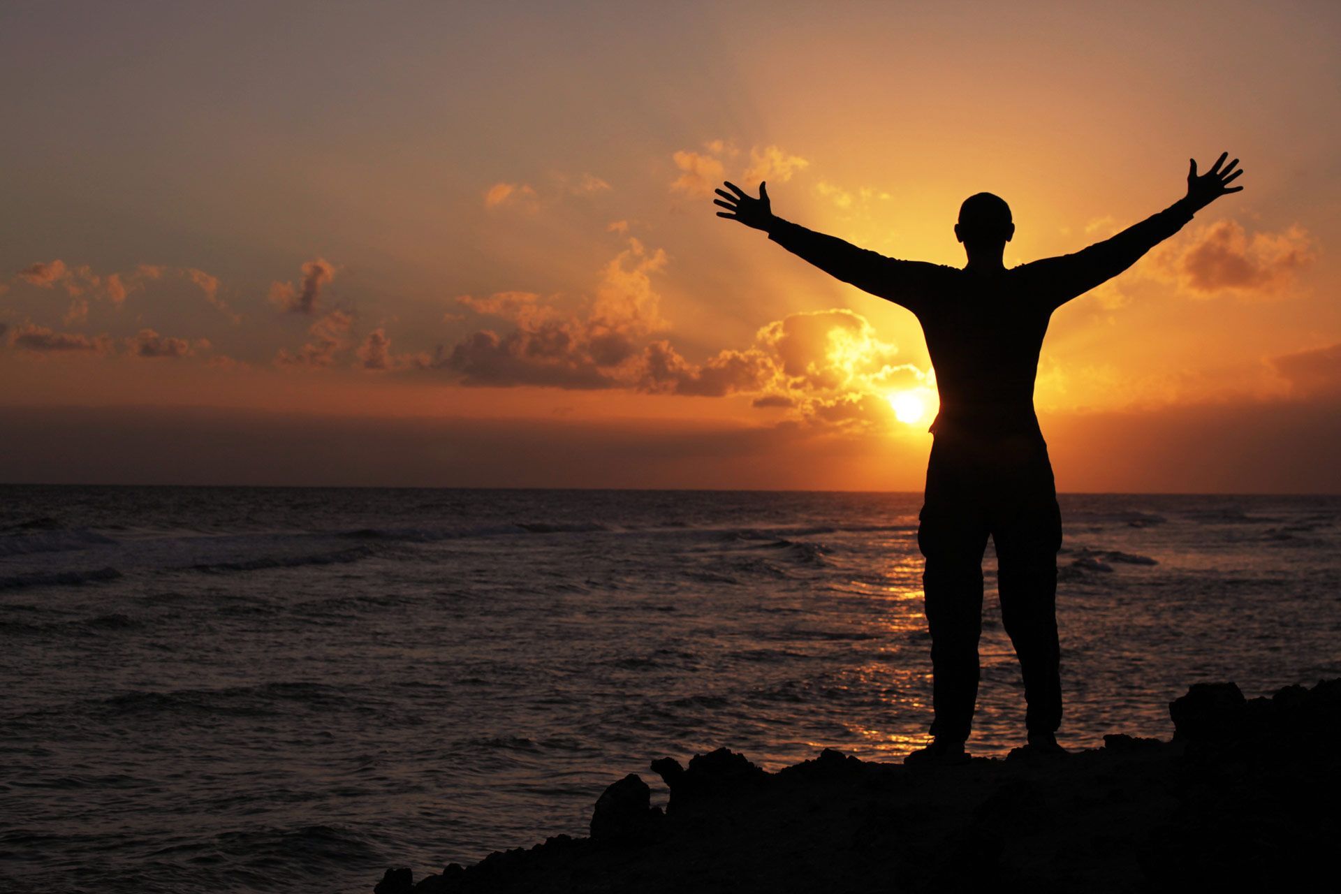 Silhouette of a person with arms outstretched, facing sunset over ocean.