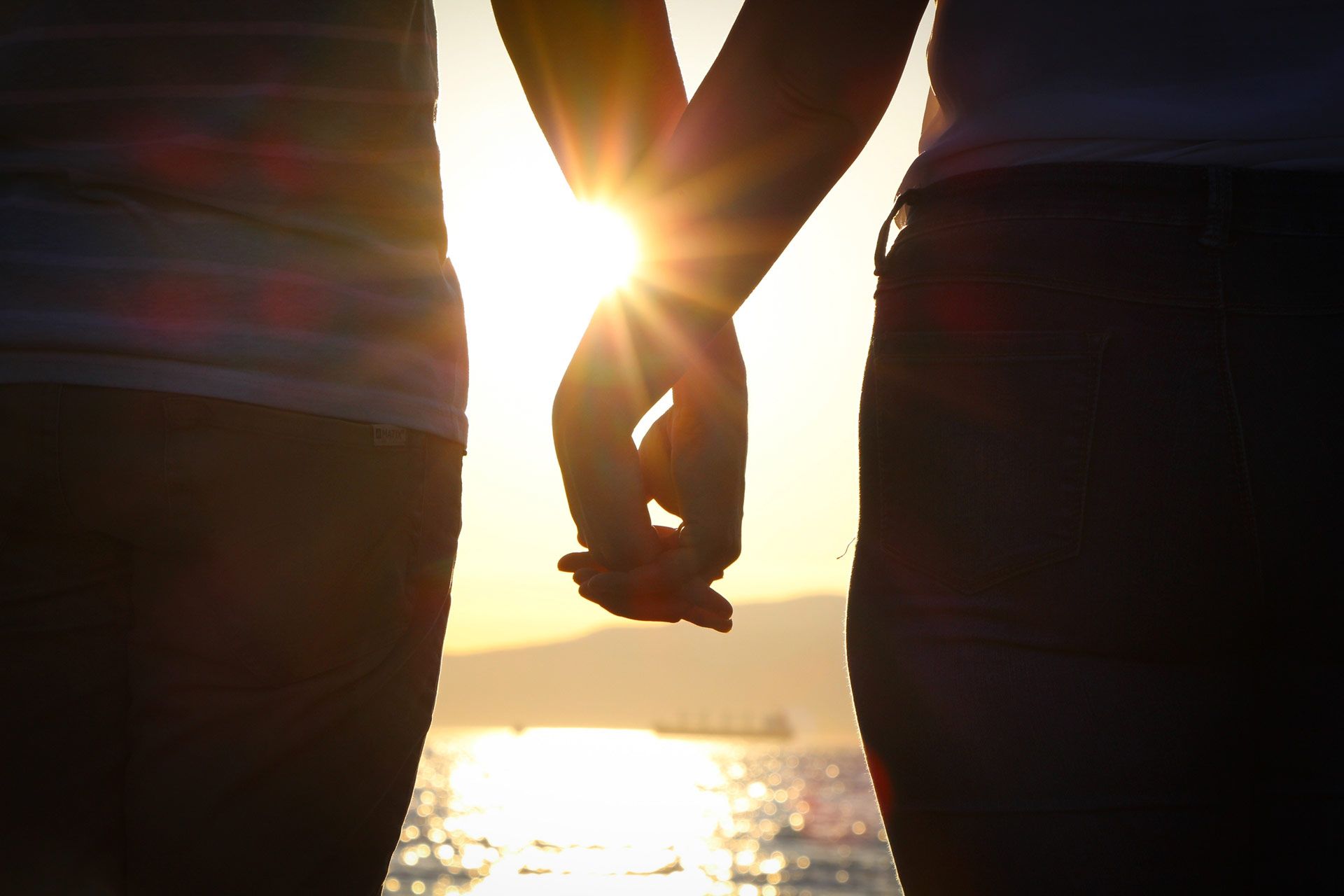 Two people holding hands, silhouetted by the setting sun over water and mountains.