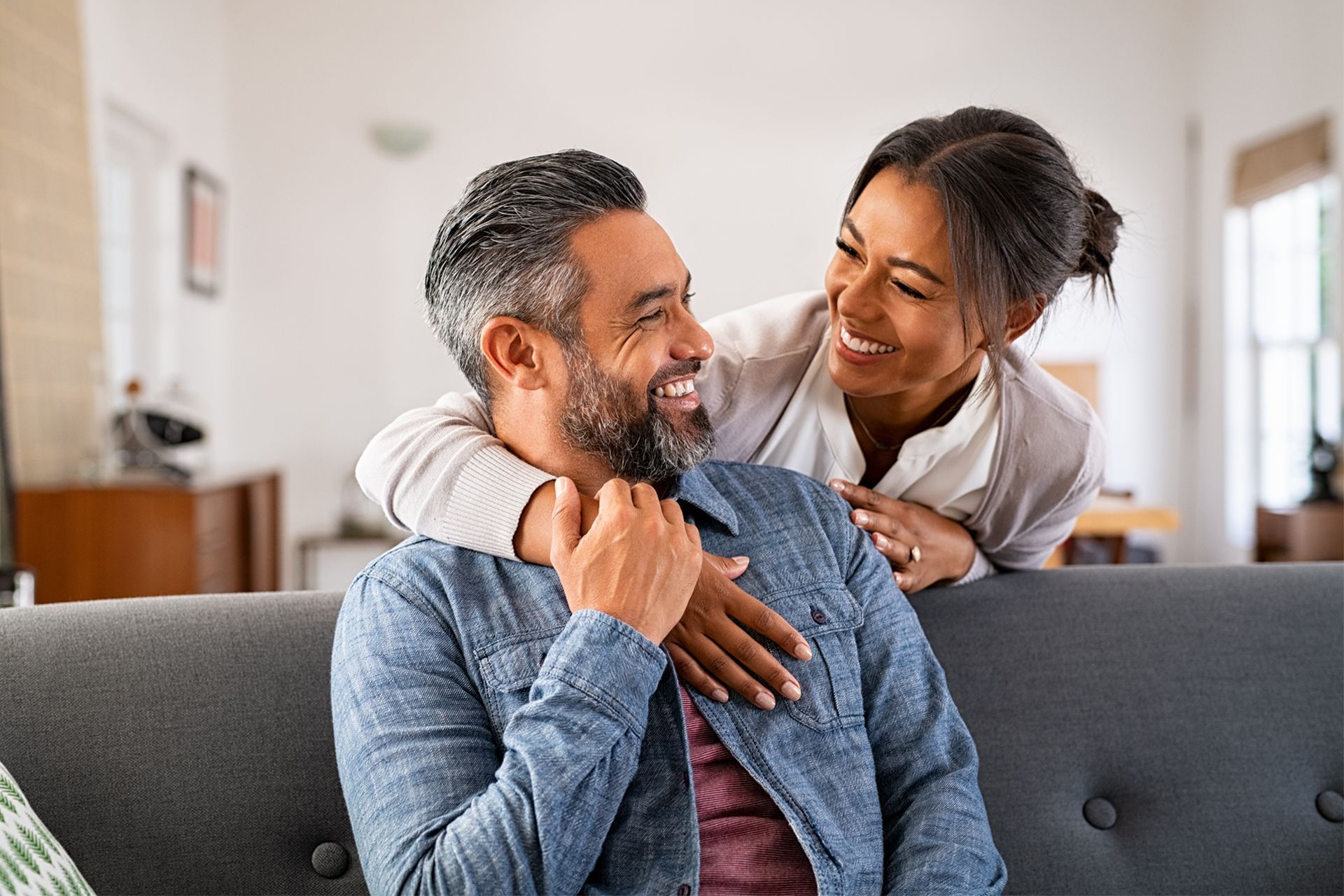 Woman embracing a man seated on a sofa.