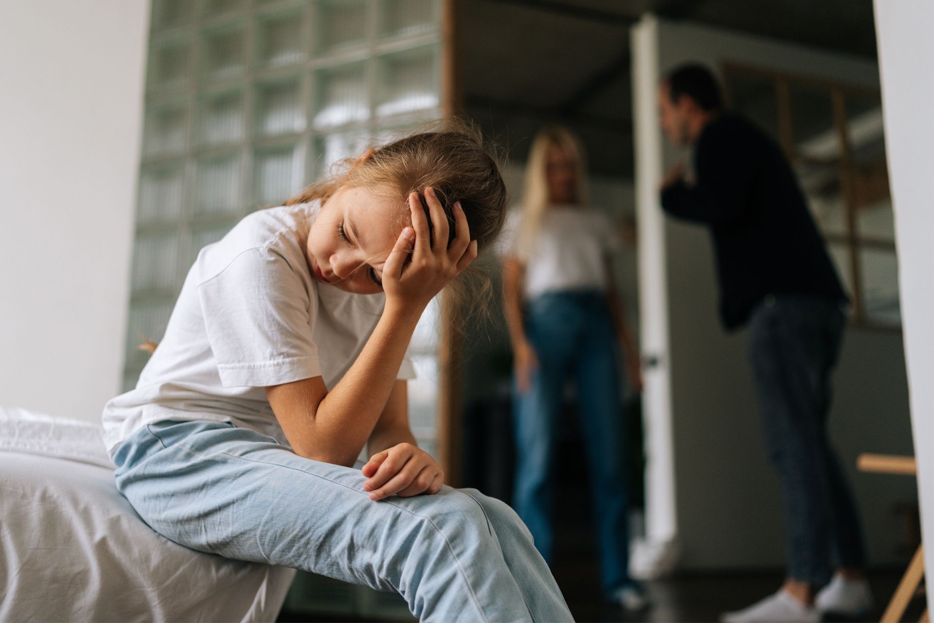 Girl with head in hand sits, parents argue in background doorway.