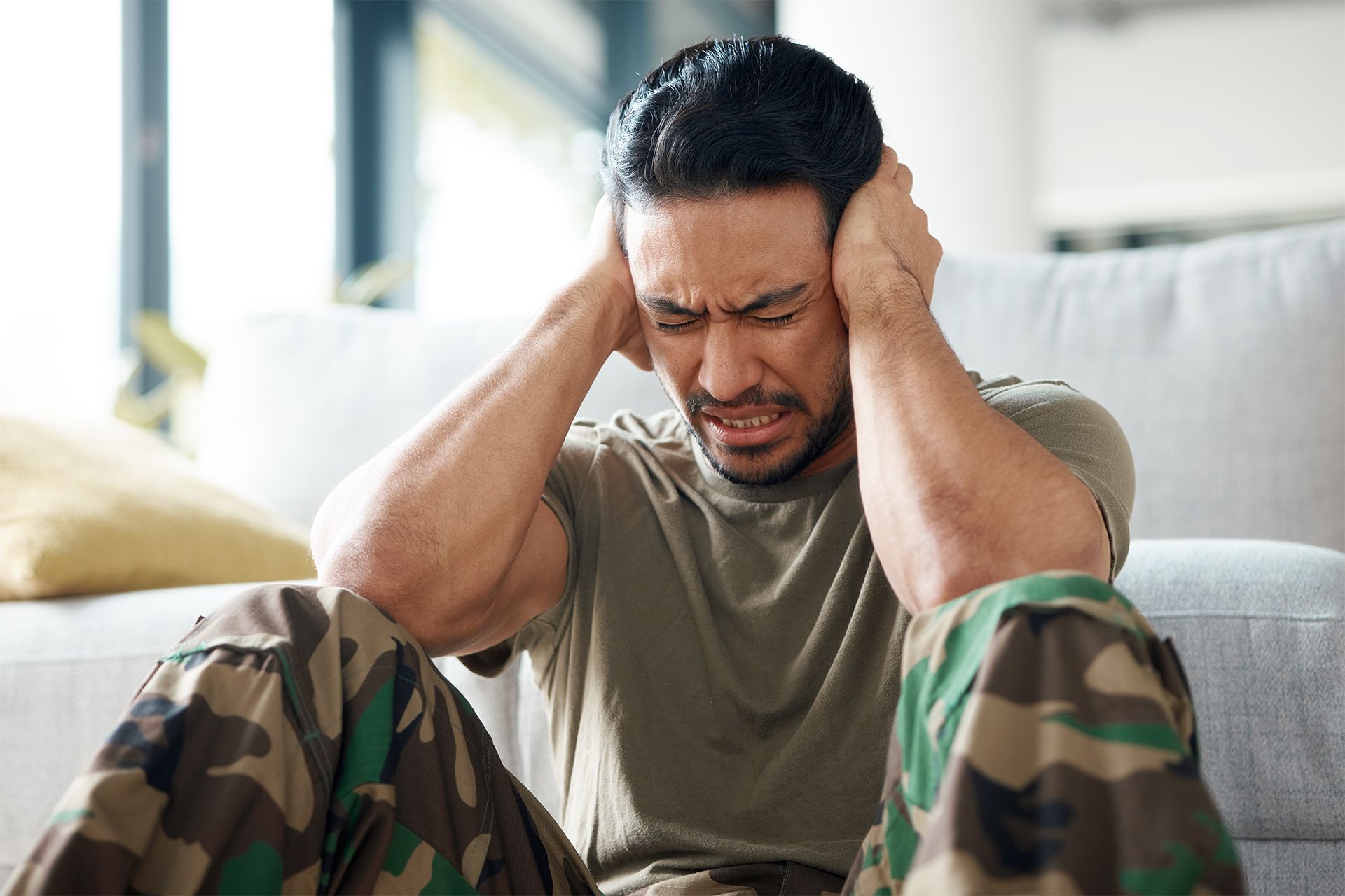 Man in camouflage pants and shirt, seated, covering ears with a pained expression.