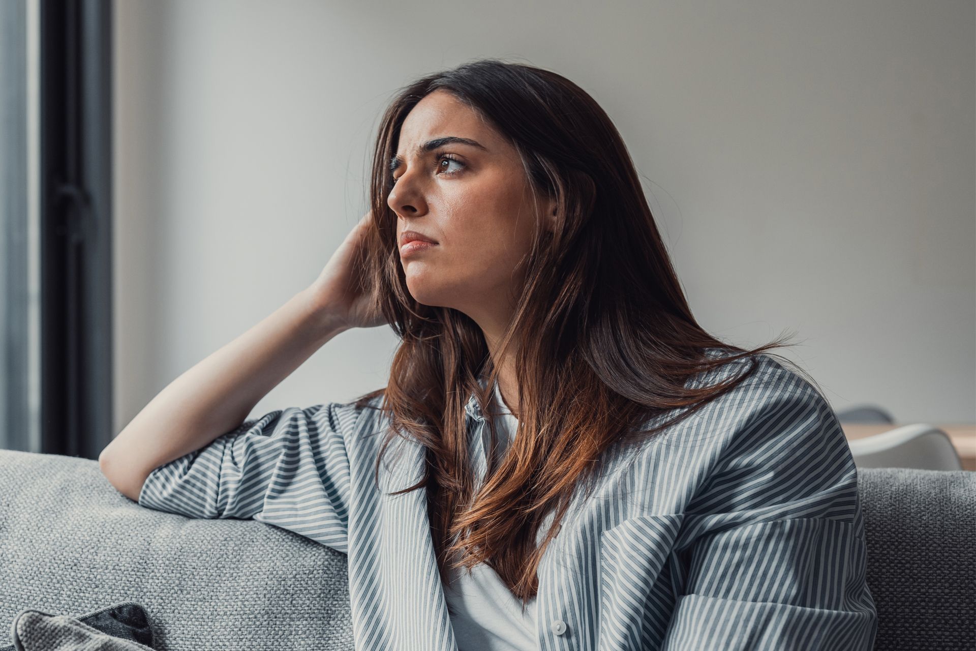 Woman sitting on a couch, looking pensive, hand to her head. Wearing a striped shirt.