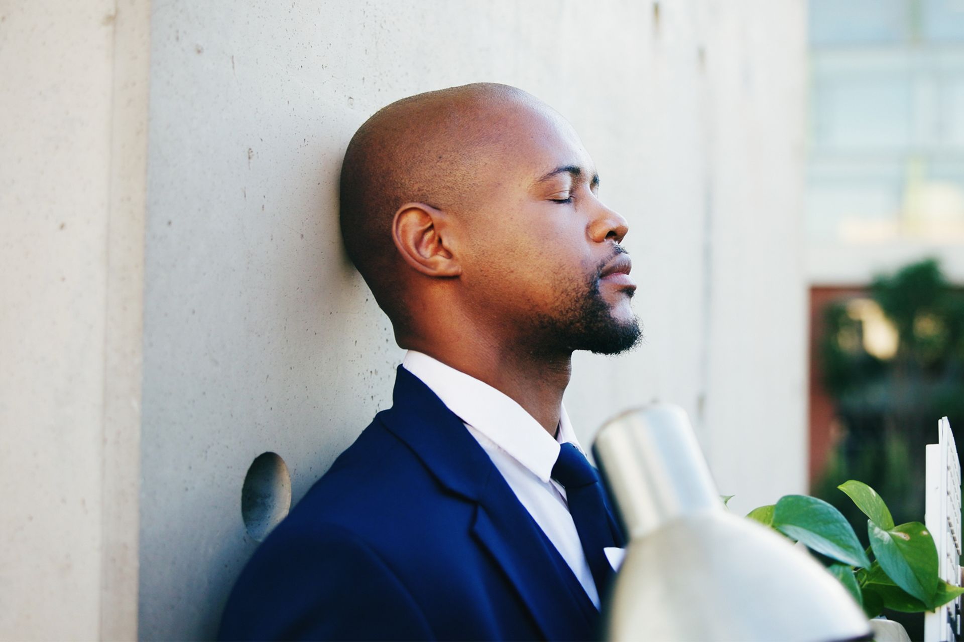 Man in blue suit, eyes closed, leaning against a wall outside.