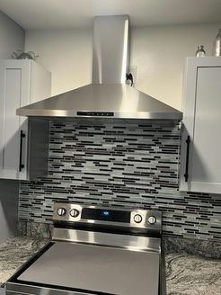 Stainless steel range hood over stove, gray and black tile backsplash, light gray cabinets.