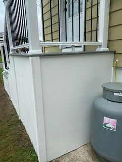 White porch wall with a gray top, next to a propane tank. Black railing and a door are in the background.