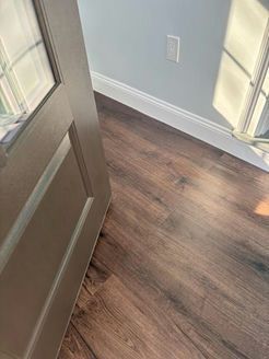 Door partially open, showing wood-look flooring, light blue wall with white trim, and a window reflecting sunlight.