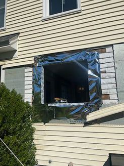Hole in house wall where window was, covered with reflective tape. Exterior view.