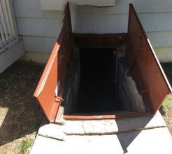 Brown cellar door open, leading to a dark, underground space on the side of a house.