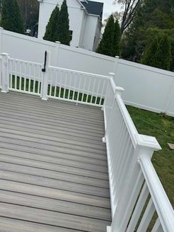 Gray deck with white railing, fence, and a glimpse of a house in the background. Green grass surrounding.