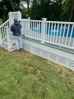 Person painting a white deck railing next to a swimming pool on a sunny day.