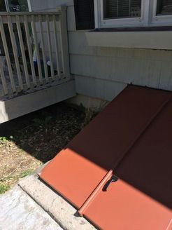 Brown metal basement door next to a wooden porch. Weeds and dirt in the space. Gray siding and concrete.