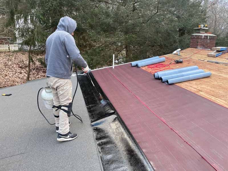 Roofer using a torch on a flat roof. Red and black roofing material visible. Outdoor setting with trees.