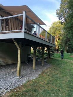 Elevated wooden deck with tan awning, black railings, supported by posts. A person walks on the grass nearby.