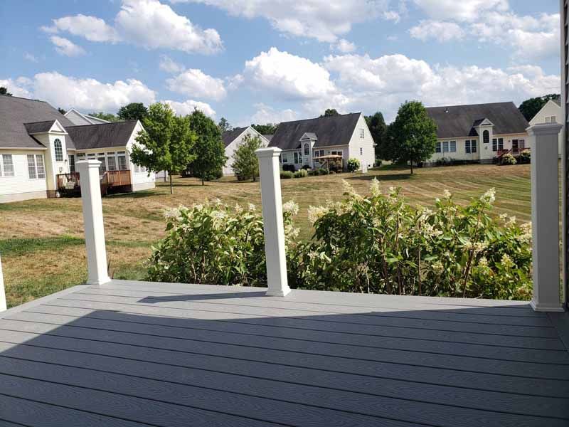 View from a gray deck overlooking a grassy yard with houses and puffy clouds in a blue sky.